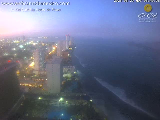 A hazy twilight view shows a coastal city at night with high-rise buildings and a dark ocean with waves visible near the shore.