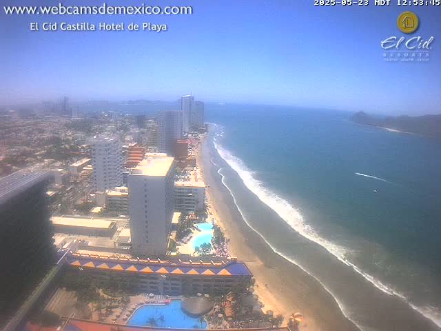 An aerial view shows a beach with gently lapping waves under a clear, sunny sky, backed by high-rise hotels and a city.