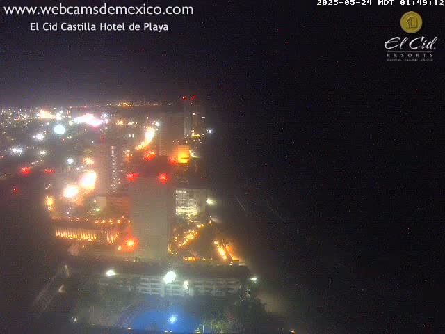 A nighttime aerial view of a brightly lit coastal city with a large hotel complex, under a clear, dark sky.