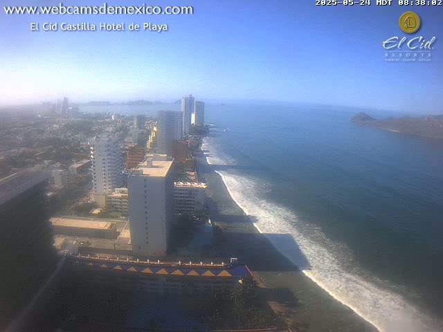 A coastal city with high-rise buildings and a sandy beach with gentle waves under a clear, sunny sky.