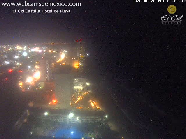 A nighttime aerial view of a coastal city shows brightly lit buildings and streets extending to a dark ocean under a clear sky.