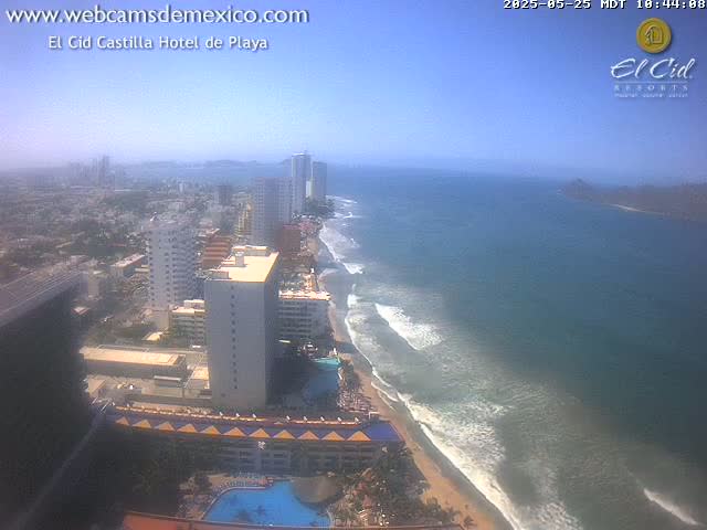 An aerial view shows a hazy beach with gentle waves, hotels, and high-rise buildings along a coastline.
