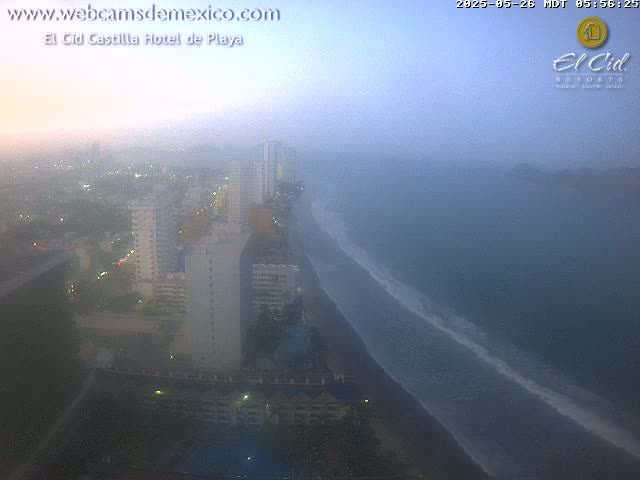 A hazy coastal city at dawn is seen from a high vantage point, showing a beach with waves and numerous buildings.