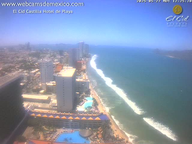 A high-angle, sunny view shows a beach with breaking waves and a cityscape of tall buildings along the coast.