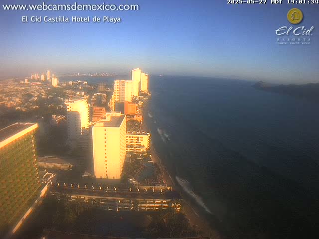 A coastal city at sunset, with numerous high-rise buildings and a dark blue ocean, under a clear sky.