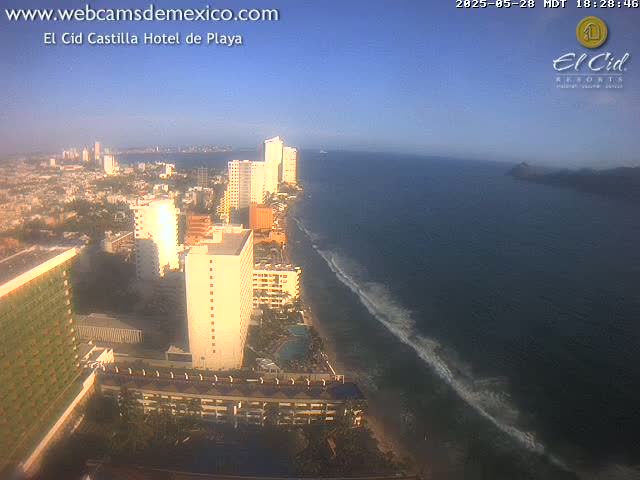 A high-angle, long shot shows a coastal city with many high-rise hotels and buildings along a sandy beach with gentle waves under a clear blue sky.