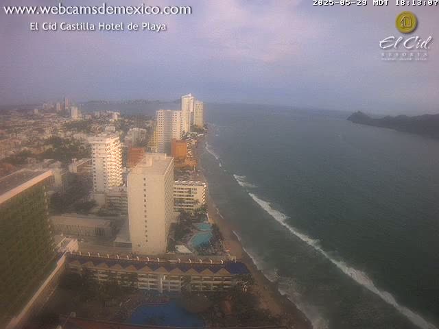 An aerial view shows a hazy coastal city with high-rise hotels along a sandy beach and ocean with gentle waves.