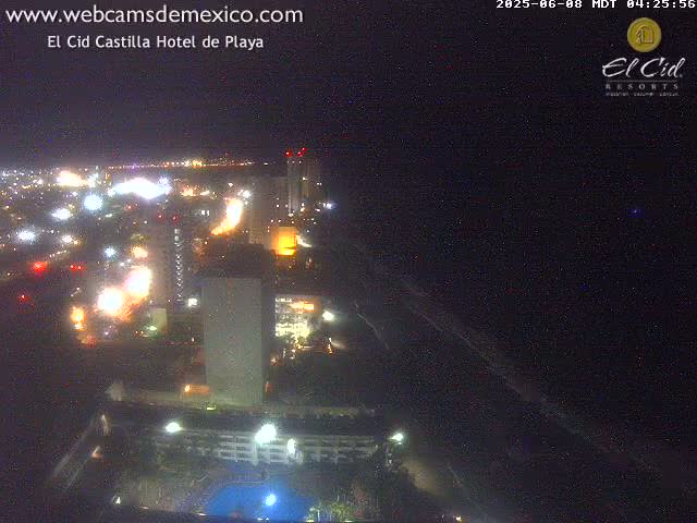 A nighttime aerial view of a city coastline showing brightly lit buildings and streets next to a dark beach under a clear sky.