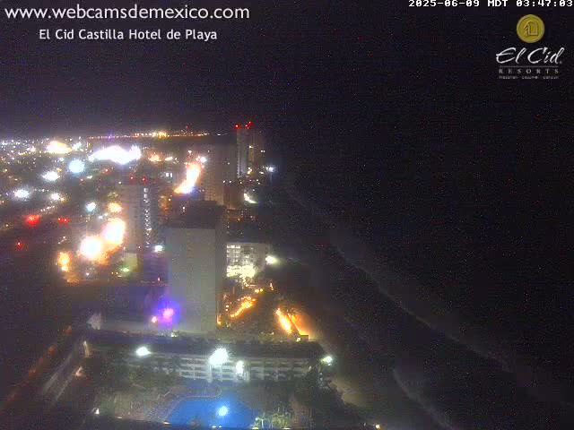 A nighttime aerial view of a brightly lit coastal city with a dark ocean and gently rolling waves.