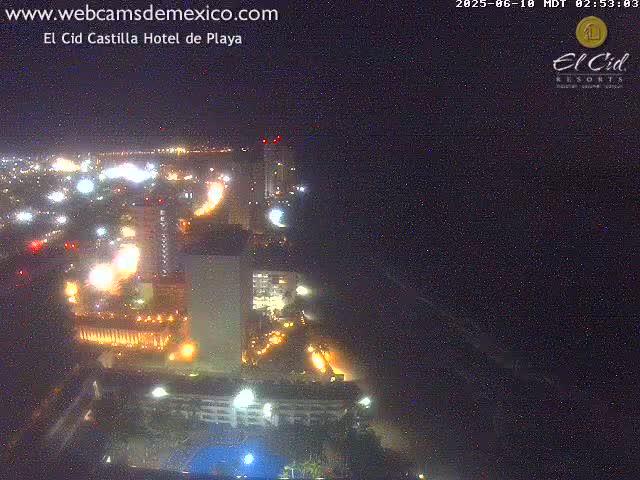 A nighttime, aerial view of a brightly lit coastal city with a dark ocean in the foreground.