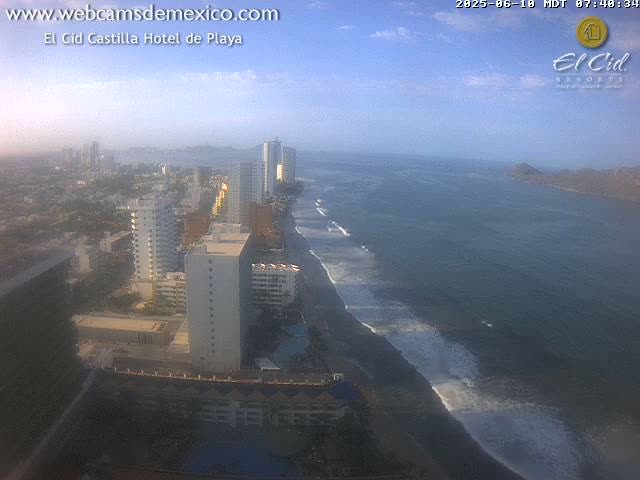 A coastal city with high-rise buildings and a dark-sand beach with light waves under a partly cloudy sky.