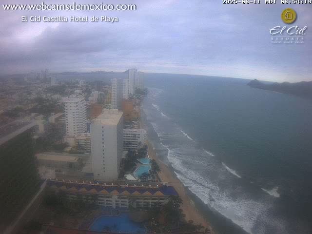A coastal city scene on an overcast day shows a beach with waves, hotels, and buildings stretching into the distance.