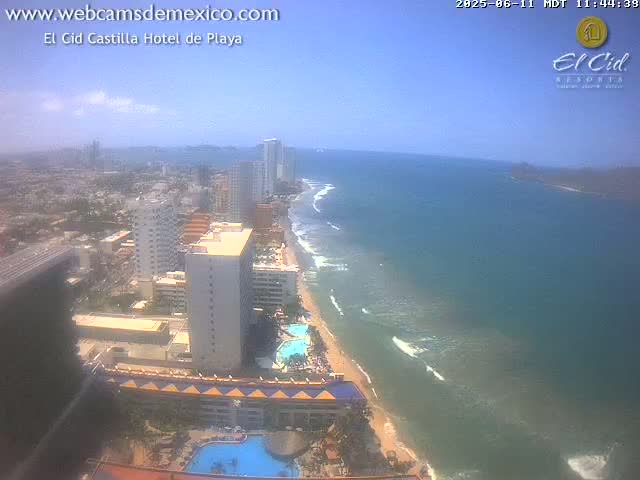 A high-angle, long shot on a sunny day shows a beach with calm ocean waves, a shoreline with hotels and buildings, and a city in the background.