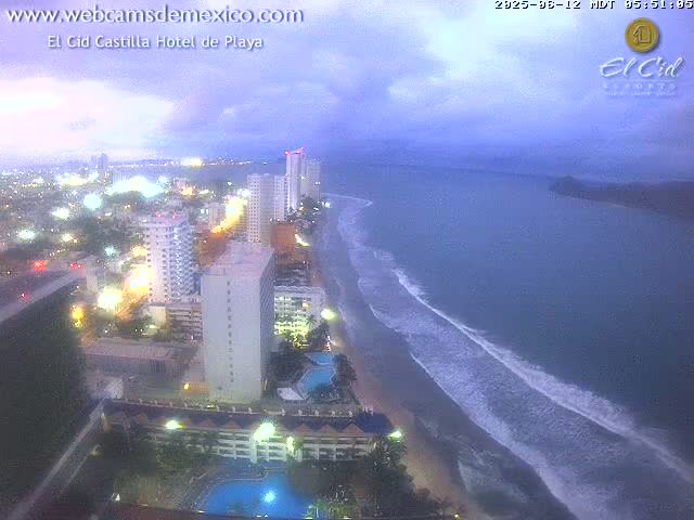 An aerial view at dusk shows a city skyline next to a beach with gentle waves under a cloudy sky.