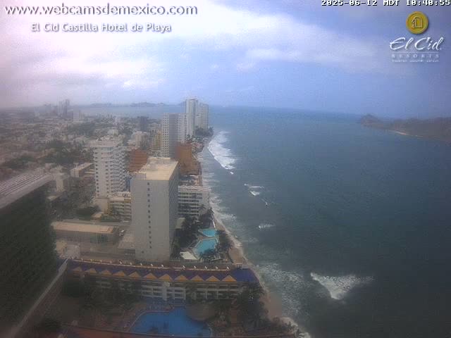A high-angle, hazy view shows a coastal city with hotels and buildings along a beach with light waves under a partly cloudy sky.