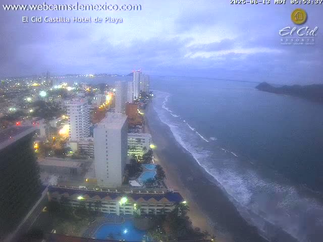 An overcast evening view of a beach with waves lapping the shore, featuring a coastal city with numerous high-rise buildings illuminated at night.