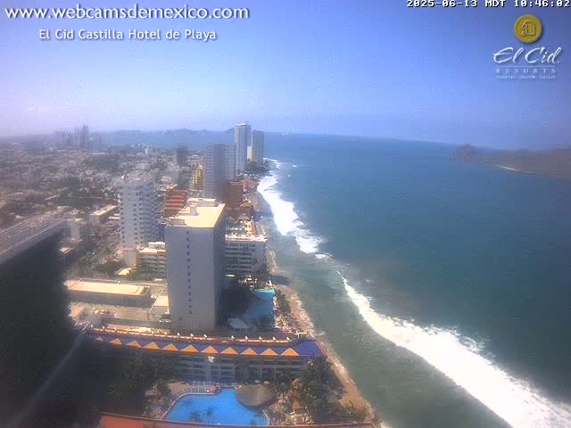A high-angle view on a clear, sunny day shows a coastline with hotels and buildings, a sandy beach, and ocean waves rolling in.