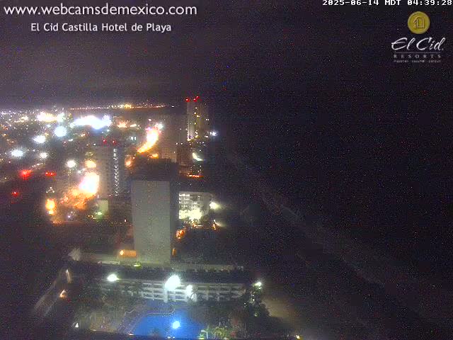 A nighttime aerial view shows a city's illuminated coastline with high-rise buildings and a swimming pool, next to a dark ocean under a clear night sky.