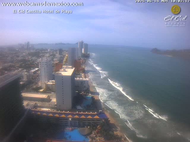 An aerial view shows a beach with waves under a hazy sky, featuring a beachfront city with high-rise hotels and buildings.
