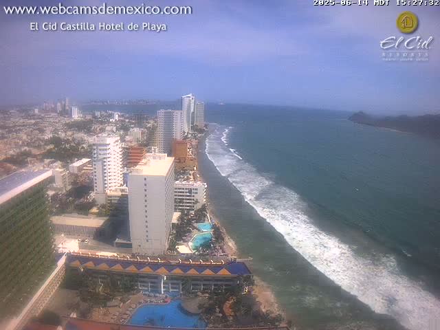 An aerial view shows a beach with waves under a hazy sky, featuring a beachfront city with high-rise hotels and buildings.