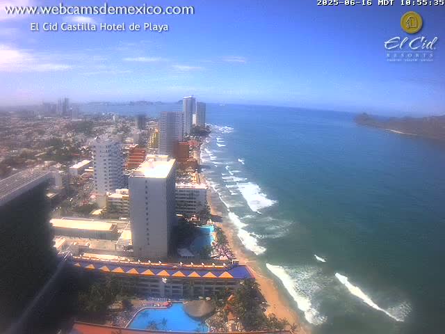 An aerial view shows a beach with gentle waves under a clear blue sky, next to a city with high-rise buildings.