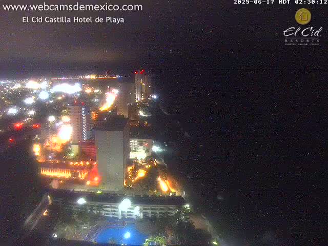 A nighttime aerial view of a city coastline showing brightly lit buildings and roads, with a dark ocean to the right.