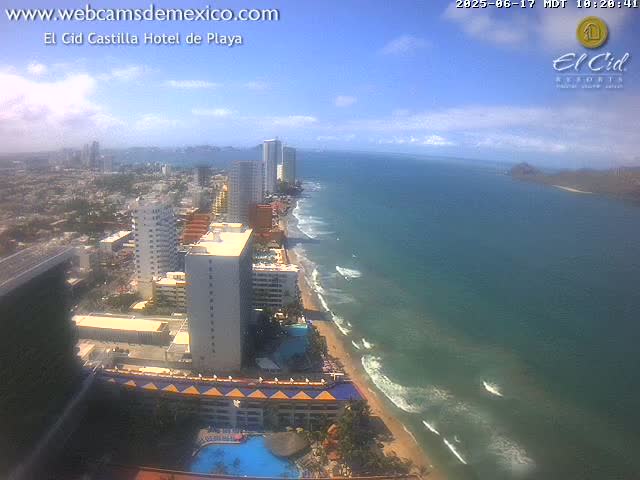 A partly cloudy day shows a coastal city with high-rise hotels and a long sandy beach with gentle waves lapping the shore.