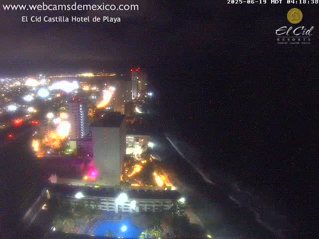 A nighttime aerial view shows a brightly lit coastal city with buildings and roads, stretching to a dark ocean with small waves.