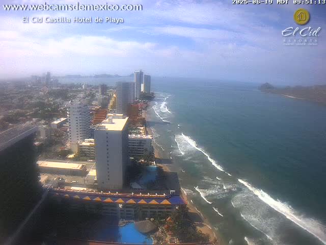 An aerial view shows a partly cloudy day over a beach town with high-rise hotels, and waves breaking on the shore.