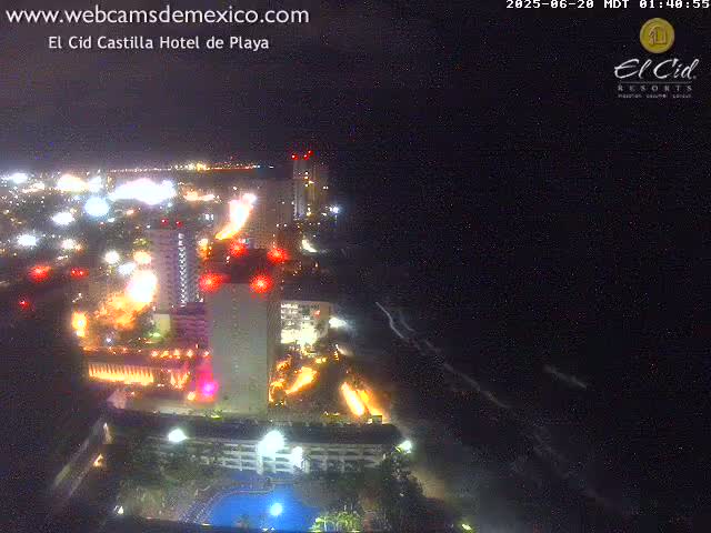 A nighttime view of a brightly lit coastal city with a hotel and ocean waves.