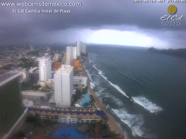 An aerial view of a coastal city under a dark, overcast sky shows several high-rise hotels and buildings along a sandy beach with waves breaking in the ocean.