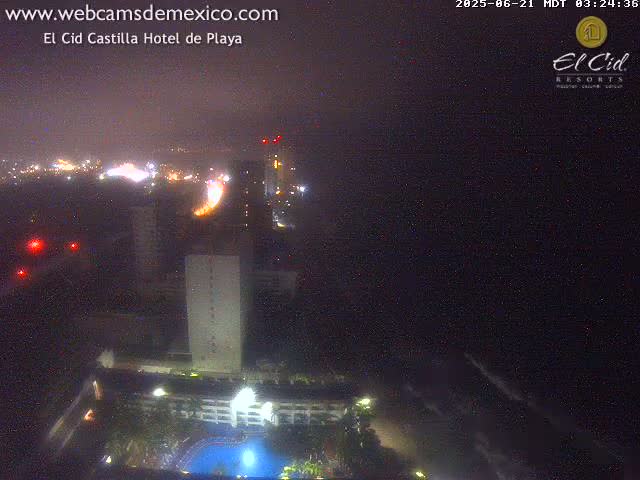 A nighttime, hazy view of a city coastline with lit buildings, a swimming pool, and dark ocean.