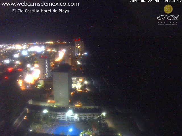A nighttime aerial view shows a brightly lit coastal city with numerous buildings and a swimming pool, extending to a dark ocean under a clear sky.