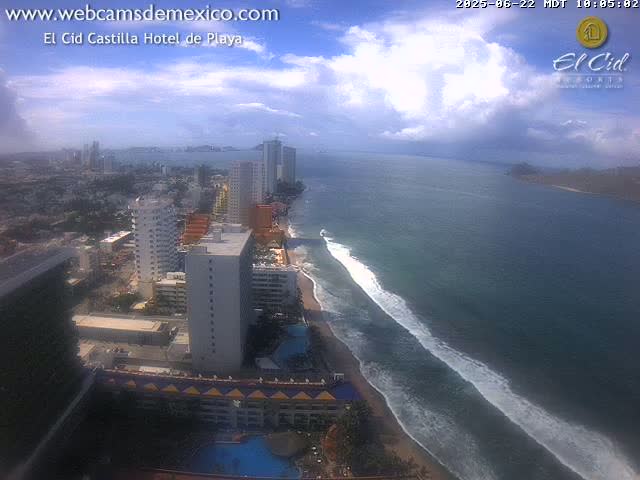 A partly cloudy day reveals a coastal city with high-rise hotels and buildings, a sandy beach with waves rolling in from a dark blue ocean.