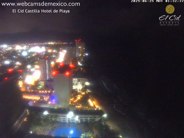 A nighttime aerial view of a brightly lit coastal city with a dark ocean and a partly cloudy sky.