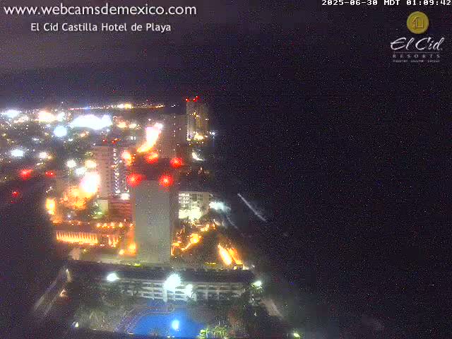 A brightly lit resort complex at night, situated near a dark ocean under a clear, starry sky.