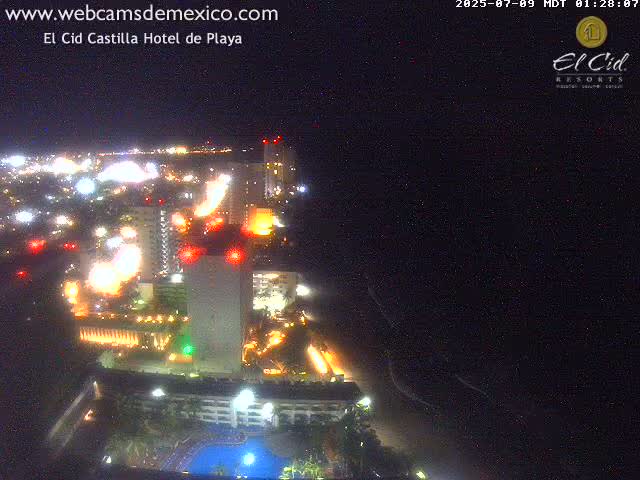 A brightly lit coastal resort town at night, viewed from above, with a dark ocean stretching to the right.