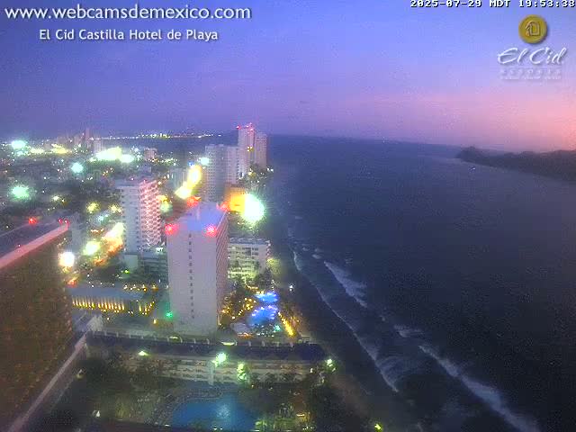 A brightly lit coastal resort town at night, viewed from above, with a dark ocean stretching to the right.