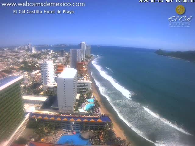 A high-angle, sunny day view shows a long sandy beach with waves rolling in towards a city skyline of high-rise buildings and hotels.
