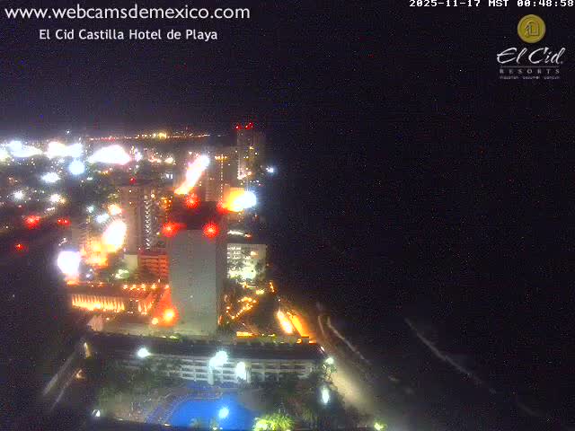 An aerial night view reveals a brightly lit coastal city with numerous buildings and roads, a discernible pool area, and a dark ocean with faint breaking waves under a clear sky.