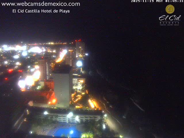 An elevated night view reveals a brightly lit coastal city with numerous buildings and a beach stretching along the dark ocean, featuring a visible swimming pool in the foreground, all under clear nighttime conditions.