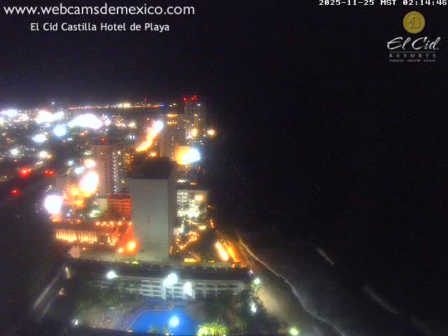 A vibrant nighttime aerial view captures a brightly lit city with numerous buildings and a hotel pool sprawling along a dark coastline, with the ocean visible under a clear sky.