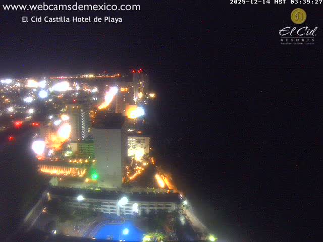 A vibrant nighttime aerial view captures a brightly lit city with numerous buildings and a hotel pool sprawling along a dark coastline, with the ocean visible under a clear sky.