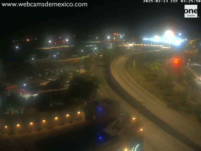 A nighttime, high-angle view shows a highway curving through a landscape with sparse vegetation and buildings, lit by streetlights.