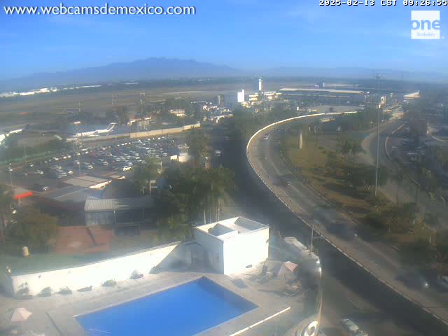 An elevated view on a clear day shows an airport with numerous parked cars, a highway curving around a building with a swimming pool, and mountains in the distance.