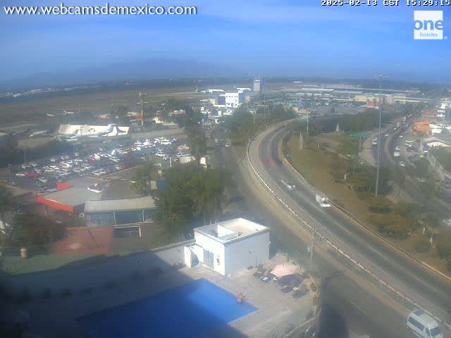 A high-angle, long shot on a clear day shows a highway curving past a hotel with a swimming pool,  a large parking lot, and an airport in the distance.