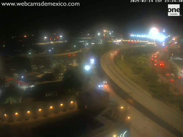 A nighttime aerial view of a highway with moderate traffic, passing near brightly lit buildings and an airport under clear skies.