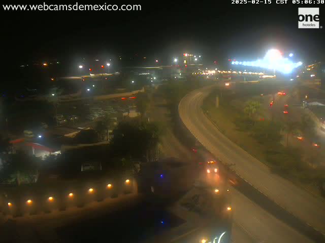 A nighttime, high-angle view shows a highway curving through a landscape with sparse vegetation, buildings, and airport lights in the distance.