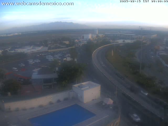 A hazy sunrise reveals a highway curving past a parking lot and a building with a pool, all situated near a flat expanse of land that seems to be an airport, with mountains faintly visible in the distance.