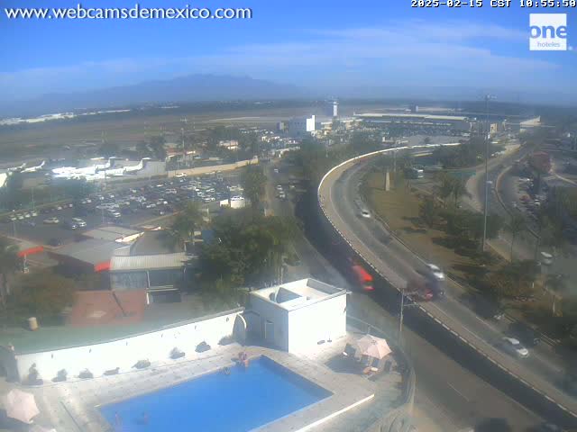 An elevated view shows a highway curving around an airport with parked planes and cars, a swimming pool, and a clear, sunny sky.